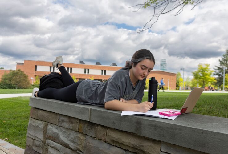student lying down on a wall along Harpur Quad working on homework with a laptop