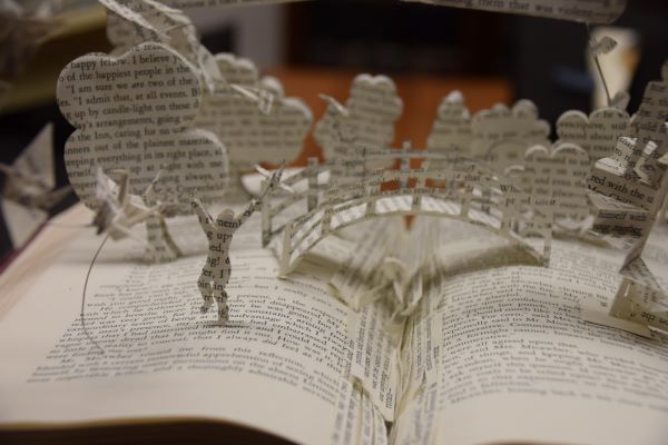 Close up photograph of a book sculpture of a scene of trees, a bridge and a person made from printed pages springing from an open book.