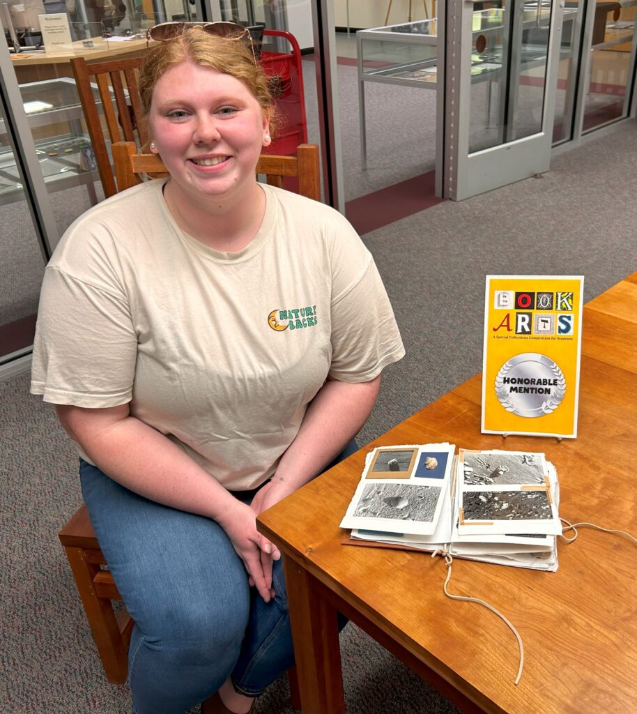 Hayley Eggleston sitting at a table beside her multimedia book "Exploration of the Seashore," Honorable Mention 2024, with certificate placed on the table above the book.