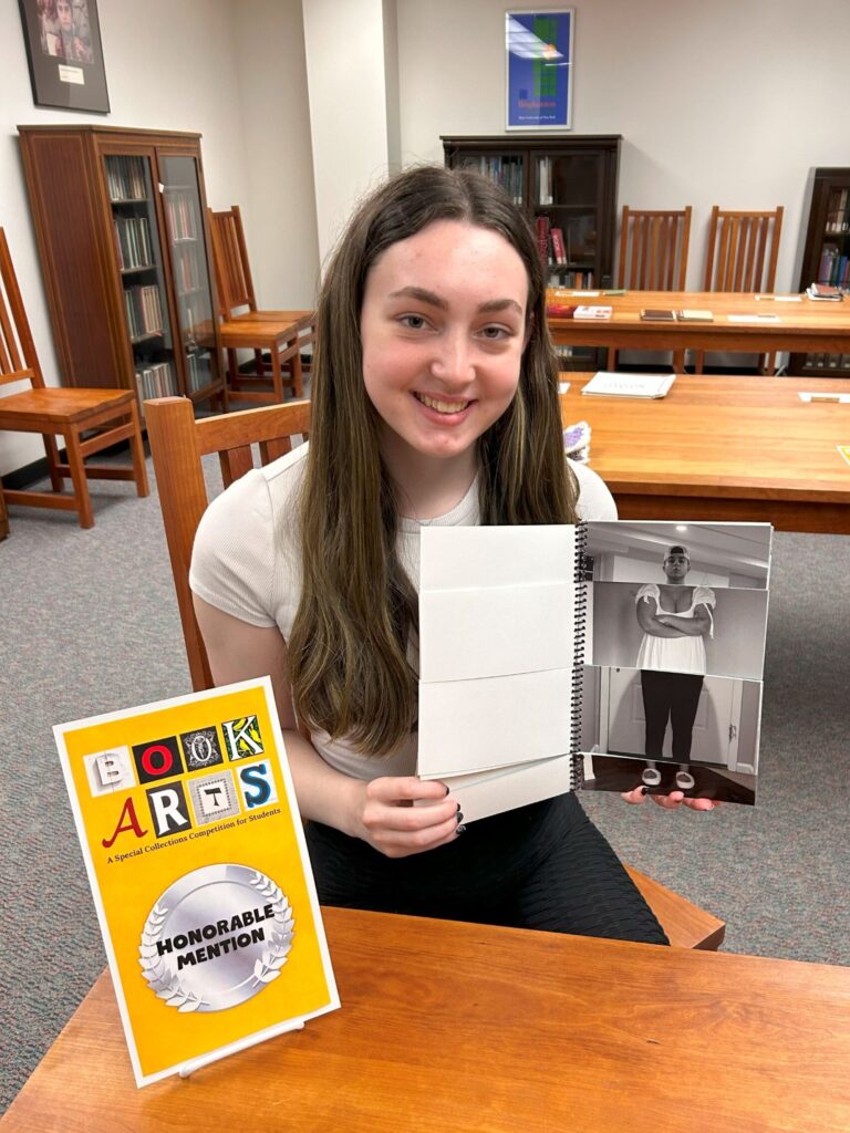 Photo of Rachel Knee holding her flap book "Indecisive Attire," Honorable Mention 2024, with certificate sitting on the table in front of her.