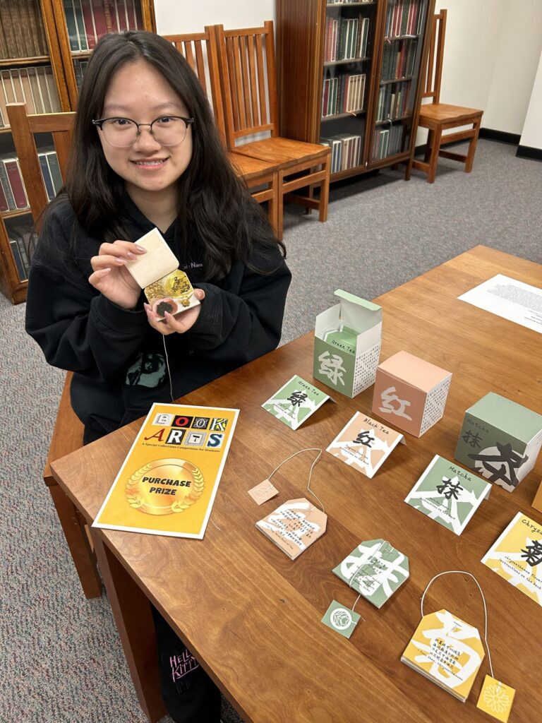 Photo of student Sammy Feng holding one of the tea bag booklets that are part of her larger work called "Cha". She is seated at a table that has the rest of her book arts piece of boxes and booklets resembling tea packaging and tea bags. A sign that says "Book Arts Purchase Prize" is on the table next to the work.