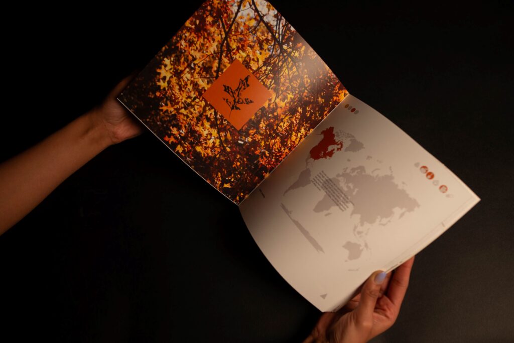 Two hands holding open a two page spread of a book by Agamya Rao, which shows on the left side a photograph of tree branches in fall colors and a print of a leaf and on the right side a graphic map of the world highlighting areas in North America where the tree species is found.