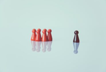 Multiple anthropomorphic, red game pieces stand in a group, separated from a single black game piece