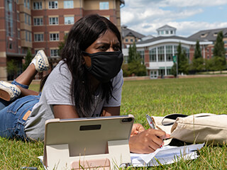 A female student lays in the grass studying while wearing a mask
