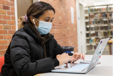 Library patron sits at a common study table next to book shelves working on her computer while wearing a mask