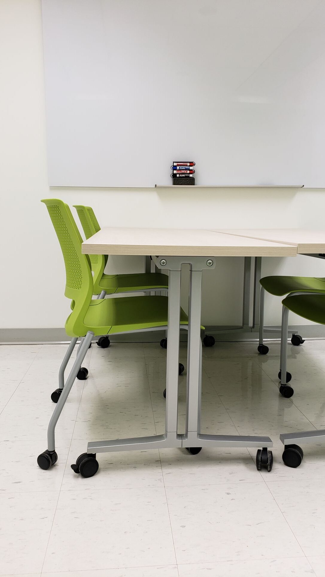 Green chairs on wheels under a table also on wheel in front of a glass board 