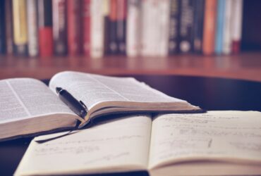 two open books on a table with a fountain pen resting in the crease of one of the books. A bookshelf can be seen in the distant background.