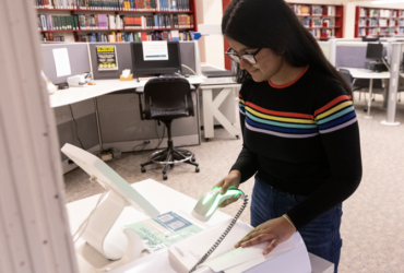 student in black sweater with a rainbow design stands at the self-scan book check out at the Science Library scanning a book's barcode with a handheld scanner.