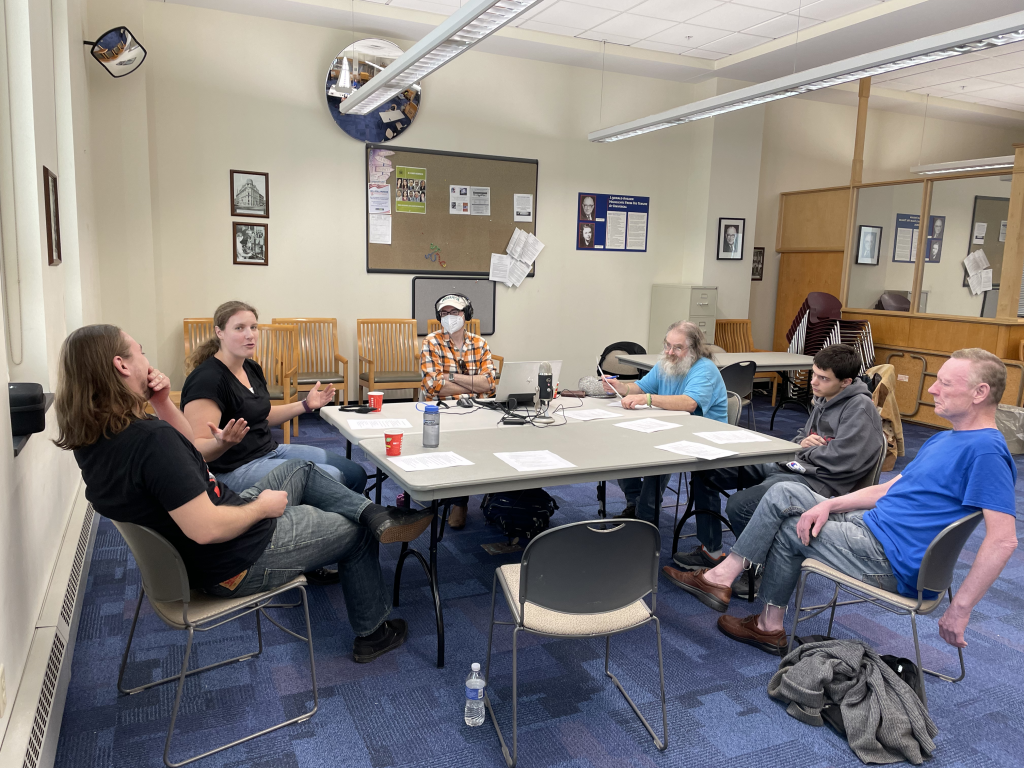 6 people sit around a table in a meeting room engaged in conversation around a microphone 