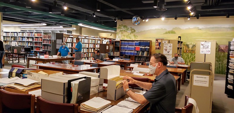 rows of books and tables with archival file folders and people sifting through them