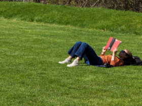 Student laying in a sunny, grassy area reading a book