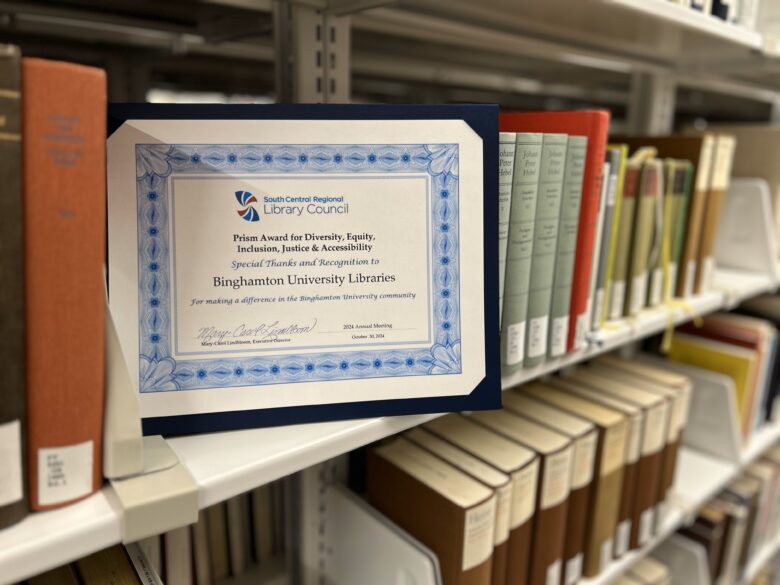 photo of the PRISM Award certificate on a bookshelf amongst various books.