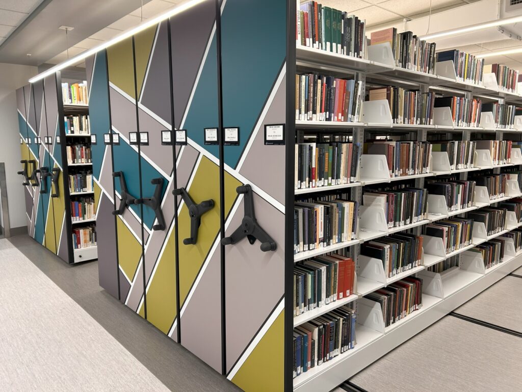 Photo of the condensed shelves located on the third floor of Bartle Library. The end of the shelves feature brightly colored geometric shapes that combine to create a mosaic pattern.