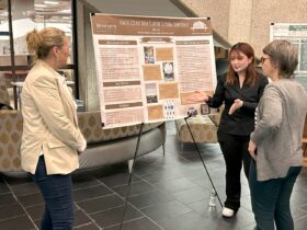 student motions towards her poster on display showing it to two library faculty during the Libraries Research Showcase in Bartle Library.