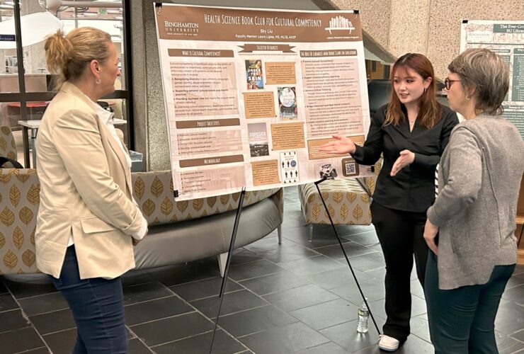 student motions towards her poster on display showing it to two library faculty during the Libraries Research Showcase in Bartle Library.