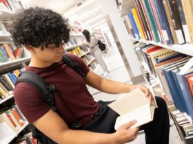 student sits and reads a book between bookshelves of books.