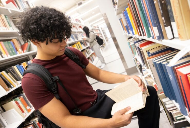 student sits and reads a book between bookshelves of books.
