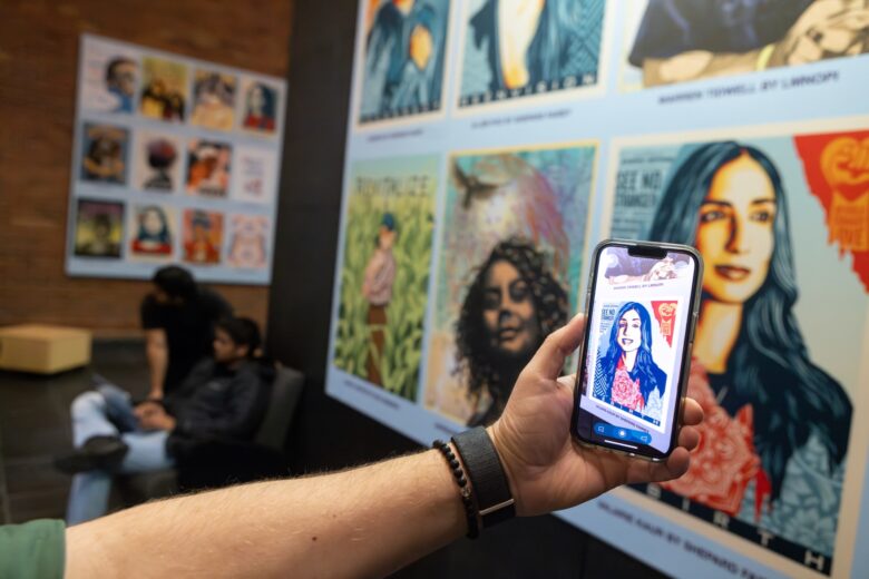 Outstretched arm holds a phone towards a collection of art portraits displayed on the first floor of Bartle Library showcasing the Augmented Reality app that animates the image.