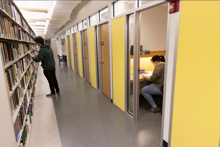 Student sits at a desk in a study Carrel with the door open while another student browses a nearby bookshelf.