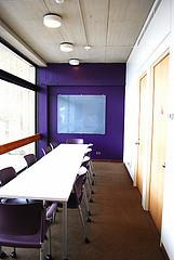 A conference-style room with a long table, chairs, and a purple accent-wall on the far end.