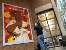 Student stands in front of a featured art piece in Bartle Library with phone outstretched to utilize the Amplifier augmented reality app. The portrait, “Patrisse Cullors” by Noa Denmon features a black woman with her hand over her heart surrounded by birds and flowers with the words “Reimagine” over her head.