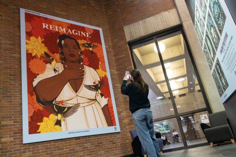 Student stands in front of a featured art piece in Bartle Library with phone outstretched to utilize the Amplifier augmented reality app. The portrait, “Patrisse Cullors” by Noa Denmon features a black woman with her hand over her heart surrounded by birds and flowers with the words “Reimagine” over her head.