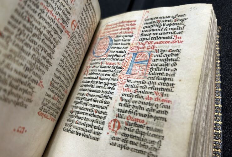 The breviary sits open-faced on a table displaying ornate script in latin. The letters feature black ink as well as larger, artistic letters in red and blue ink.