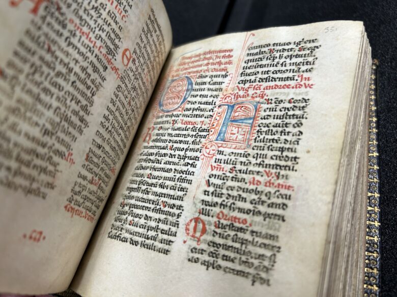 The breviary sits open-faced on a table displaying ornate script in latin. The letters feature black ink as well as larger, artistic letters in red and blue ink.