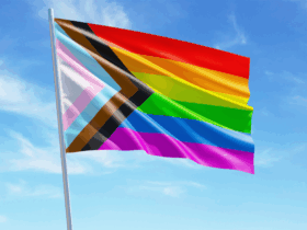 The Progress Pride flag, featuring stripes of red, orange, yellow, green, blue, and purple, alongside diagonal stripes of black, brown, light blue, pink, and white, waves against a bright blue sky with scattered clouds.