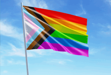 The Progress Pride flag, featuring stripes of red, orange, yellow, green, blue, and purple, alongside diagonal stripes of black, brown, light blue, pink, and white, waves against a bright blue sky with scattered clouds.