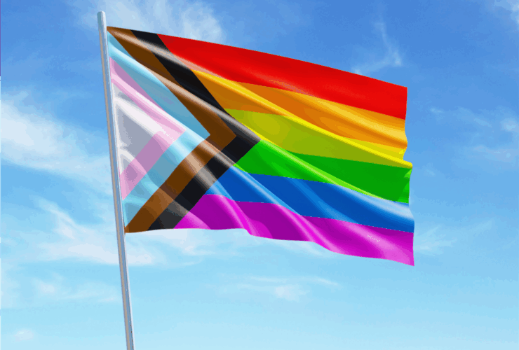 The Progress Pride flag, featuring stripes of red, orange, yellow, green, blue, and purple, alongside diagonal stripes of black, brown, light blue, pink, and white, waves against a bright blue sky with scattered clouds.